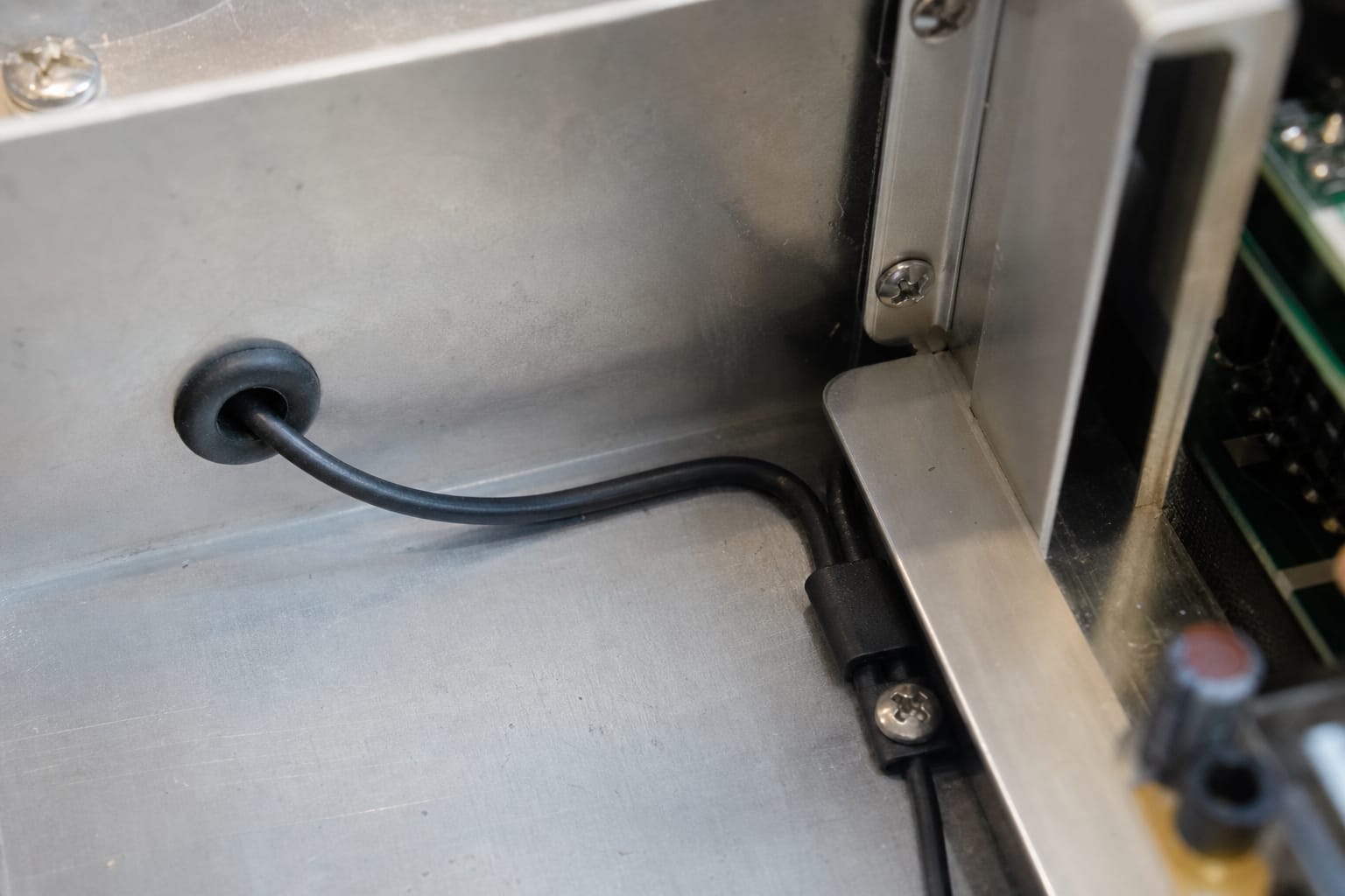 A thin black-jacketed thermosensitive sensor cable routed through a rubber grommet in the wall of a brushed-aluminium equipment enclosure and clamped around a sharp inside corner, the bend held close to its minimum bend radius, with cross-head fixing screws and the edge of a green circuit board visible alongside — the kind of tight in-housing routing where a cable's mechanical envelope, not its electrical spec, decides whether it fits