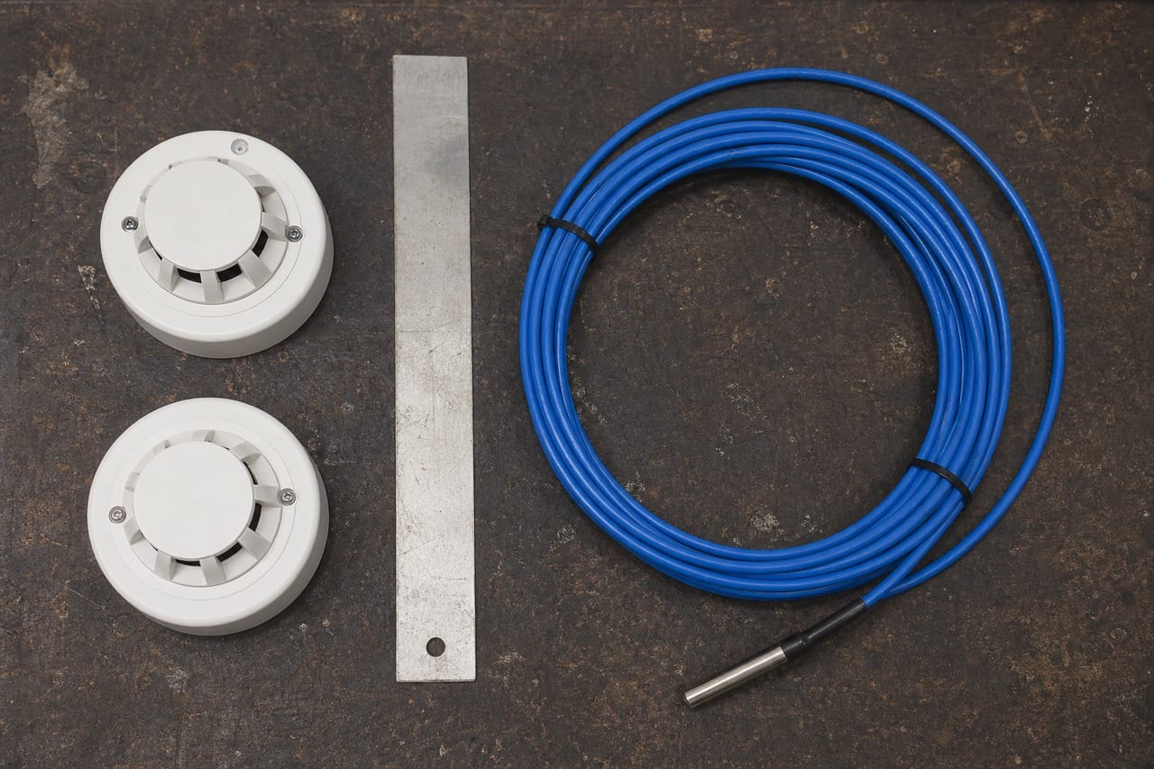 Overhead view on a dark workbench comparing two heat-detection methods — two white round spot (point) heat detector heads on the left, a steel ruler in the centre for scale, and a coiled blue linear heat detection (LHD) cable with a stainless-steel end fitting on the right
